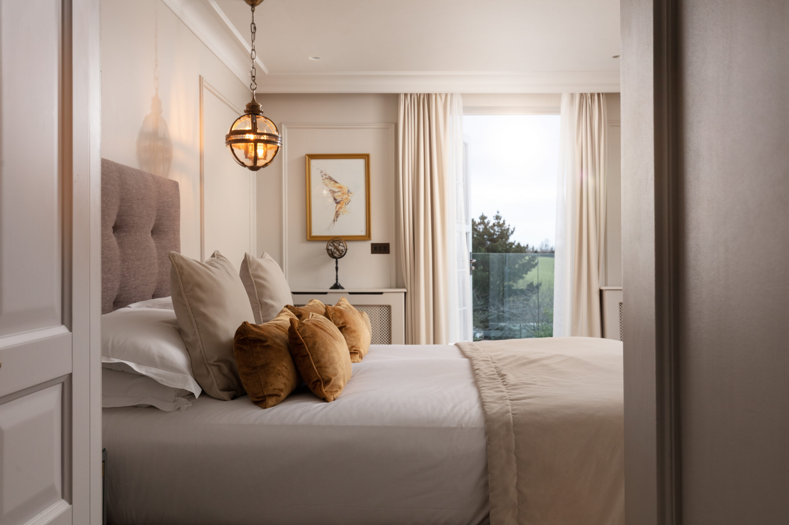 Lympstone Manor suite with a view to the balcony and out to the estate with bed, cushions, light and artwork on the wall.